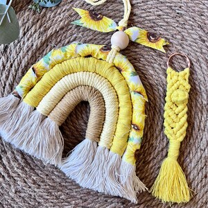 May include: A yellow, white, and brown macrame rainbow wall hanging with a sunflower fabric accent. The rainbow has white fringe and is attached to a yellow cord with a wooden bead. A matching yellow macrame keychain with a sunflower accent is also included.