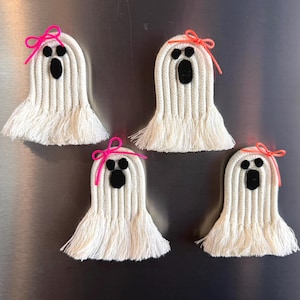 May include: Four ghost-shaped decorations made of white coiled rope with frayed fringe bottoms. Each ghost features two black felt eyes and an oval black felt mouth. Two ghosts have bright pink bows, and two have neon orange bows.