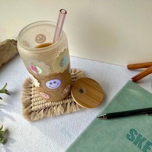 May include: A clear glass tumbler with a pink straw, filled with a light brown beverage, decorated with smiley face and flower stickers. The tumbler sits on a beige macrame coaster, next to a journal, pen, and lit candle.