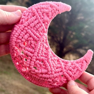 May include: A pink crocheted crescent moon wall hanging. The moon is made of a soft, textured yarn and has a delicate, intricate design.