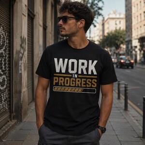 Pode incluir: Camiseta preta com as palavras "WORK IN PROGRESS" em letras brancas e douradas. O design inclui um gráfico de barra de progresso. A camiseta está sendo usada ao ar livre.