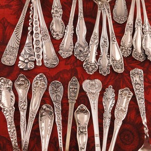 May include: A collection of vintage silver teaspoons with ornate designs. The spoons have intricate floral and scrollwork patterns, and some feature a small, raised figure at the end of the handle.