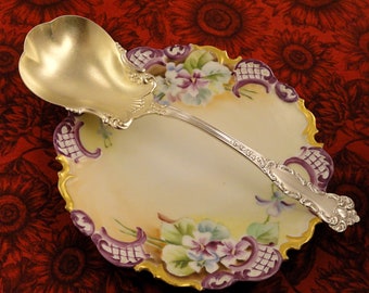 May include: A silver serving spoon with an ornate handle and a gold-plated bowl, resting on a decorative porcelain plate with a floral design in shades of purple, yellow, and green.