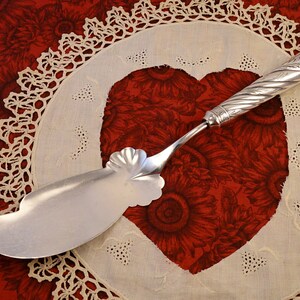 May include: A silver cake server with an ornate handle and a scalloped edge, resting on a white crocheted doily with a heart-shaped cutout.