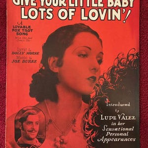 May include: A vintage sheet music cover with a red background. The title is "Give Your Little Baby Lots of Lovin'!". The cover features a black and white photo of a woman with dark hair and a necklace. The text "Introduced by Lupe Velez in her Sensational Personal Appearances" is at the bottom of the cover.