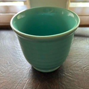 May include: A solid turquoise ceramic cup with a smooth, glossy finish and a rounded rim. The exterior features subtle, horizontal ribbed detailing that wraps around the body of the vessel, creating a simple, uniform texture throughout the piece.