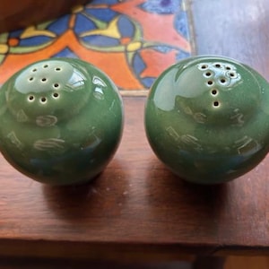 May include: Two round, green ceramic salt and pepper shakers. Each shaker has multiple holes on top for dispensing. The shakers are on a wooden surface, with a colorful tile design visible in the background.