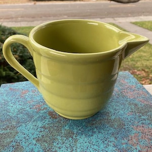 May include: A green ceramic pitcher with a wide mouth and a handle. The pitcher has a smooth, glossy finish and a slightly rounded shape.