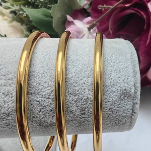 Peut inclure: Trois bracelets dorés avec une finition lisse et polie. Les bracelets sont présentés sur une surface grise et veloutée, avec un arrangement floral flou en arrière-plan. Les bracelets sont un bijou classique.