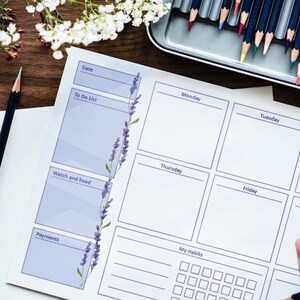 May include: A weekly planner with lavender accents, featuring sections for dates, to-do lists, and habits. The planner is on a wooden surface, with pencils and colored pencils nearby. The design includes space for each day of the week.