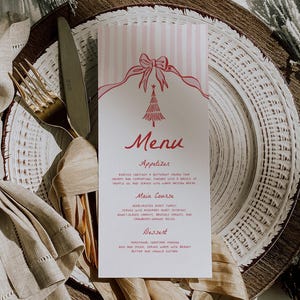 May include: A Christmas dinner table setting featuring a menu card with a pink striped design and a red Christmas tree illustration. Gold cutlery and a beige napkin are on a decorative plate.