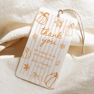 May include: A rectangular gift tag with a beige and white striped background. The tag features orange line art of pumpkins, leaves, and flowers. The words "thank you" are written in cursive, with the text "FOR COMING TO MY BIRTHDAY PARTY! Love, Amelia 10.04.2075".