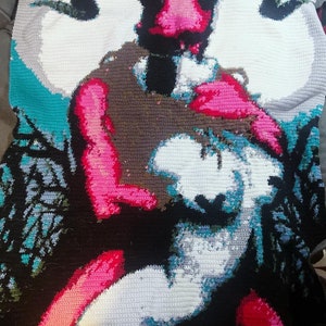 May include: A crocheted blanket featuring a colorful image of a horned figure holding a person. The blanket is made with a variety of colors, including pink, blue, green, and black.