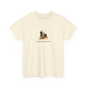 May include: A cream-coloured t-shirt with a vintage illustration of two figures in historical dress. The graphic includes the text "monitoring the situation" beneath the image in a stylized font. The shirt has a classic crew neck and is made of a soft material.