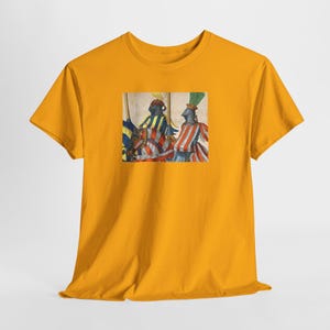 May include: Mustard yellow t-shirt featuring a rectangular graphic of two figures in medieval armor holding flags. The figures are dressed in red and white striped and blue and yellow striped clothing. The graphic is centered on the front of the shirt.