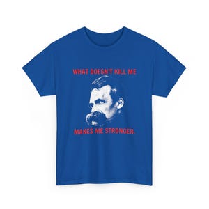 Puede incluir: Camiseta azul real con un retrato en blanco y negro de un hombre con bigote. El texto rojo "WHAT DOESN'T KILL ME MAKES ME STRONGER" está impreso arriba y abajo. La camiseta es de cuello redondo clásico.