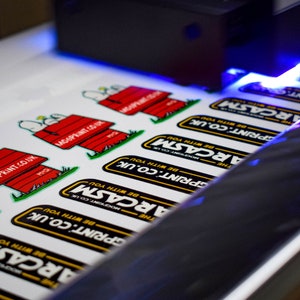 May include: Close-up of a printing machine printing stickers with the text "The ARCASM" and "MOGPRINT.CO.UK" on them. The stickers are printed on white paper and feature a cartoon dog design.