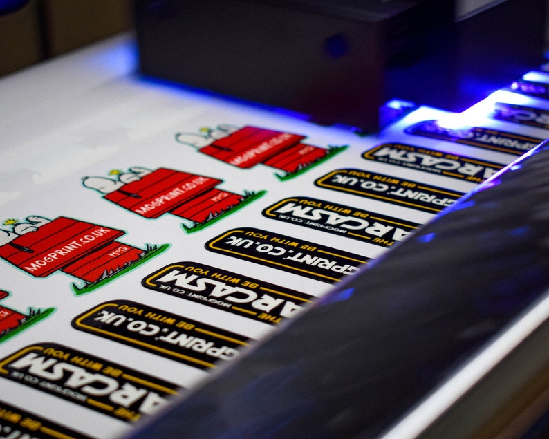May include: Close-up of a printing machine printing stickers with the text "The ARCASM" and "MOGPRINT.CO.UK" on them. The stickers are printed on white paper and feature a cartoon dog design.