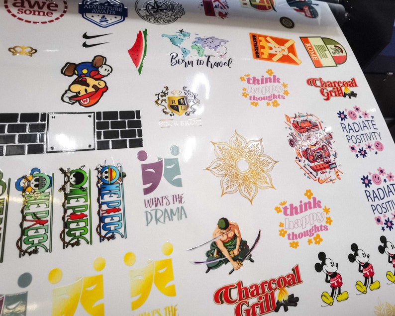 May include: A sheet of white vinyl with various colourful stickers featuring different designs and text. Some of the stickers include a cartoon character, a watermelon, a flower, a cartoon character, a brick wall, a logo, and a cartoon character with a sword. The text on the stickers includes "Born to Travel", "Think Happy Thoughts", "Charcoal Grill", "Radiate Positivity", "What's the Drama", and "Think Happy Thoughts".