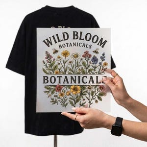 May include: A black t-shirt displayed behind a square transfer sheet featuring the text WILD BLOOM BOTANICALS. The design includes colorful illustrations of various wildflowers, green foliage, and two bees arranged in a decorative botanical pattern.