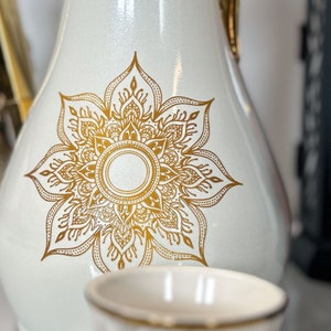 May include: White ceramic jug with a gold mandala design. The jug has a round body and a narrow neck.