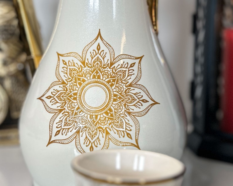 May include: White ceramic jug with a gold mandala design. The jug has a round body and a narrow neck.