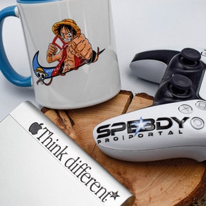 May include: A white ceramic mug with a blue handle features a colourful illustration of an anime character with a straw hat. The mug has the text "Woodprint" on the side. The mug is sitting on a wooden surface next to a white gaming controller with the text "Speedy Pro Portal" on it. A silver laptop with the text "Think different" is also visible in the image.