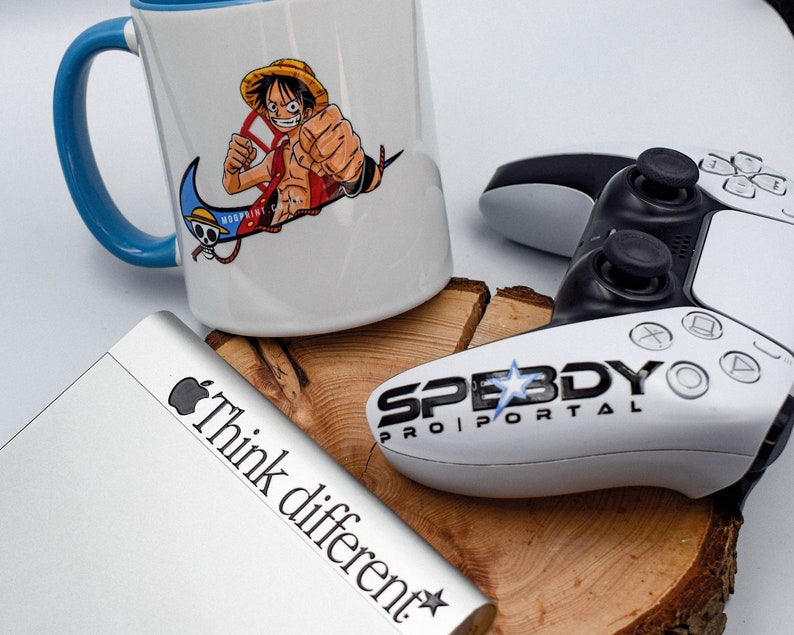 May include: A white ceramic mug with a blue handle features a colourful illustration of an anime character with a straw hat. The mug has the text "Woodprint" on the side. The mug is sitting on a wooden surface next to a white gaming controller with the text "Speedy Pro Portal" on it. A silver laptop with the text "Think different" is also visible in the image.