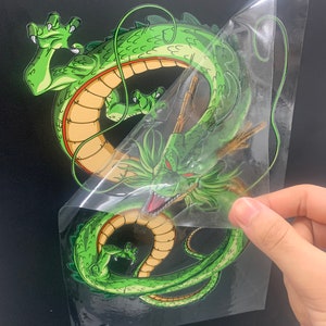 May include: Green dragon sticker with a yellow belly and a red stripe around its body. The sticker is on a clear background and is being peeled off of a black surface.