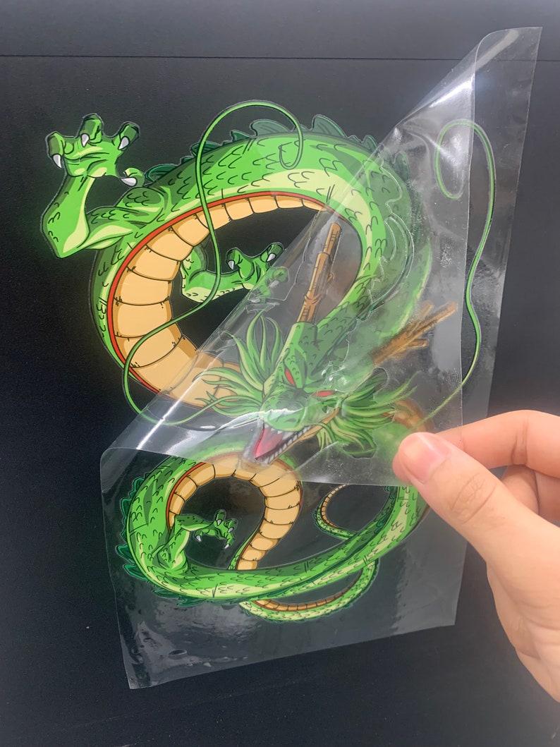 May include: Green dragon sticker with a yellow belly and a red stripe around its body. The sticker is on a clear background and is being peeled off of a black surface.