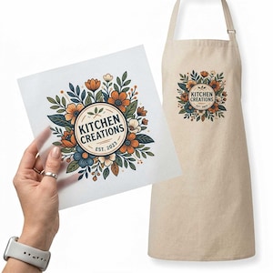 May include: A beige apron featuring a circular logo with the text KITCHEN CREATIONS EST. 2023 surrounded by orange, blue, and cream-colored flowers and green leaves. A matching graphic is shown on a square sheet of paper held in the foreground.