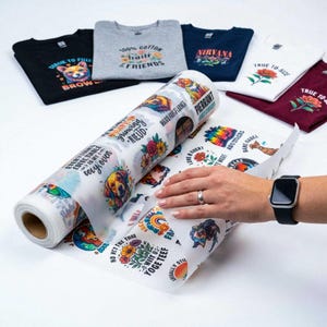 Könnte beinhalten: Eine Rolle halbtransparentes Transferpapier mit verschiedenen bunten Grafikdrucken von Hunden, Blumen und Text, platziert neben mehreren gefalteten T-Shirts in Schwarz, Grau, Marineblau, Weiß und Kastanienbraun mit passenden Druckmotiven.