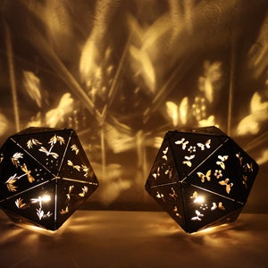 May include: Two black metal lanterns with intricate cut-out designs of butterflies and dragonflies. The lanterns are lit from within, casting warm shadows on the wall behind them.