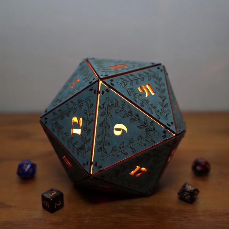 May include: A teal, illuminated D20 die-shaped lamp with gold numbers and floral detailing. The lamp is surrounded by several smaller dice on a wooden surface, creating a tabletop gaming theme.