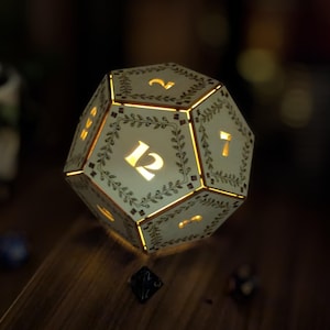 May include: A glowing, cream-colored dodecahedron die with illuminated numbers and decorative leaf patterns. The die is lit from within, casting a warm glow. The numbers 12 and 7 are visible. The overall theme is gaming and tabletop accessories.