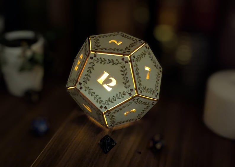 May include: A glowing, cream-colored dodecahedron die with illuminated numbers and decorative leaf patterns. The die is lit from within, casting a warm glow. The numbers 12 and 7 are visible. The overall theme is gaming and tabletop accessories.