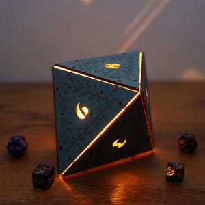May include: A teal-colored, illuminated D20 dice-shaped lamp with etched floral designs and glowing numbers. The lamp casts a warm orange light, with other dice scattered around it. A unique decorative piece for game enthusiasts.