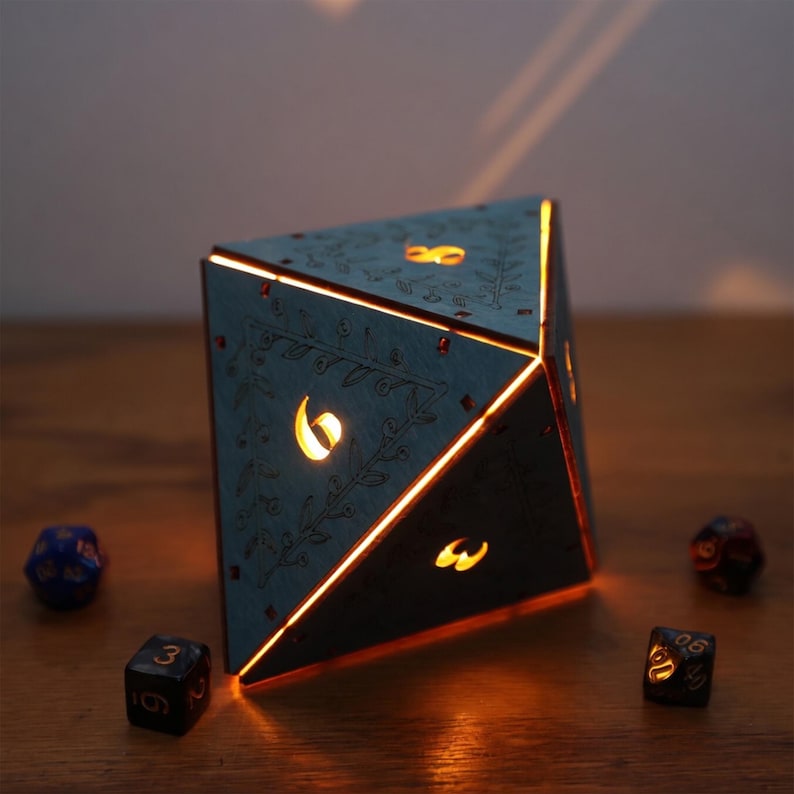 May include: A teal-colored, illuminated D20 dice-shaped lamp with etched floral designs and glowing numbers. The lamp casts a warm orange light, with other dice scattered around it. A unique decorative piece for game enthusiasts.