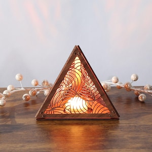 May include: A wooden, triangular-shaped lamp with intricate, geometric cutouts. The lamp emits a warm, orange glow, creating a cozy ambiance. The lamp is placed on a wooden surface, with decorative white berries in the background. The lamp is approximately 8 inches wide.