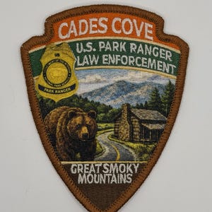 May include: A shield-shaped embroidered patch featuring the text CADES COVE, U.S. PARK RANGER LAW ENFORCEMENT, and GREAT SMOKY MOUNTAINS. The design includes a yellow park ranger badge, a brown bear, a log cabin, and a winding road in the mountains.