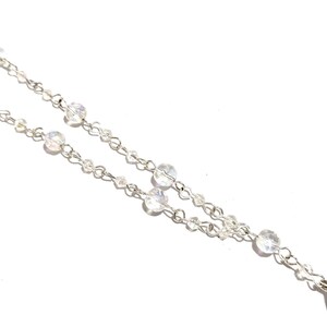 May include: Close-up of a delicate silver necklace with clear, iridescent crystal beads. The necklace features a double chain design with varying sizes of round and small beads, creating a sparkling effect against a white background. The jewelry is elegant and suitable for special occasions.