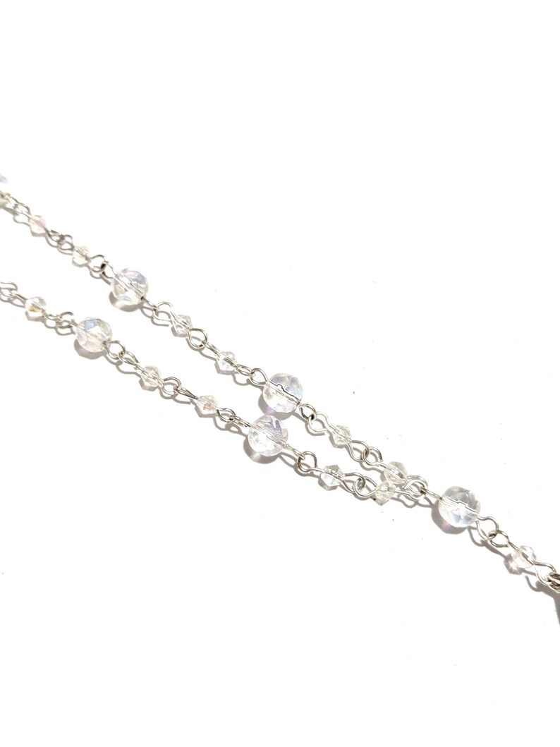 May include: Close-up of a delicate silver necklace with clear, iridescent crystal beads. The necklace features a double chain design with varying sizes of round and small beads, creating a sparkling effect against a white background. The jewelry is elegant and suitable for special occasions.