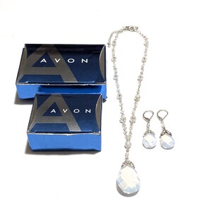 May include: A vintage jewelry set featuring a necklace and earrings with teardrop-shaped, iridescent pendants. The set is accompanied by two blue boxes with the word "AVON" printed on them. The jewelry is set against a white background.