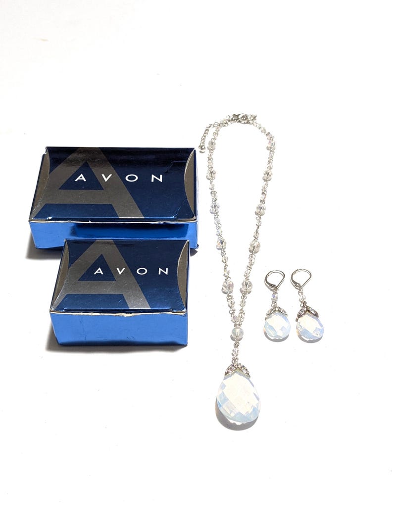 May include: A vintage jewelry set featuring a necklace and earrings with teardrop-shaped, iridescent pendants. The set is accompanied by two blue boxes with the word "AVON" printed on them. The jewelry is set against a white background.