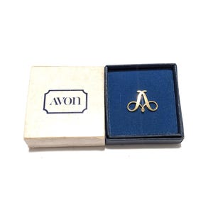 以下が含まれることがあります： 青いベルベットの裏地が付いた箱に入った、金色の文字「A」のピン。ピンは様式化された文字「A」の形をしています。箱は「AVON」のロゴが入ったクリーム色の箱の中に入っています。
