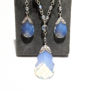 May include: A jewelry set featuring a necklace and matching earrings. The set includes a silver chain necklace with a large, faceted, teardrop-shaped pendant. The earrings also have teardrop-shaped blue gemstones. The jewelry is displayed on a gray surface.