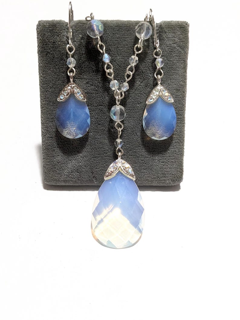May include: A jewelry set featuring a necklace and matching earrings. The set includes a silver chain necklace with a large, faceted, teardrop-shaped pendant. The earrings also have teardrop-shaped blue gemstones. The jewelry is displayed on a gray surface.