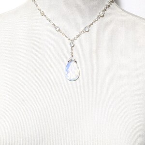 May include: A delicate silver necklace with a teardrop-shaped, iridescent pendant. The necklace features small, clear beads and a chain that drapes gracefully. The pendant is a soft, opalescent color, adding a touch of elegance.