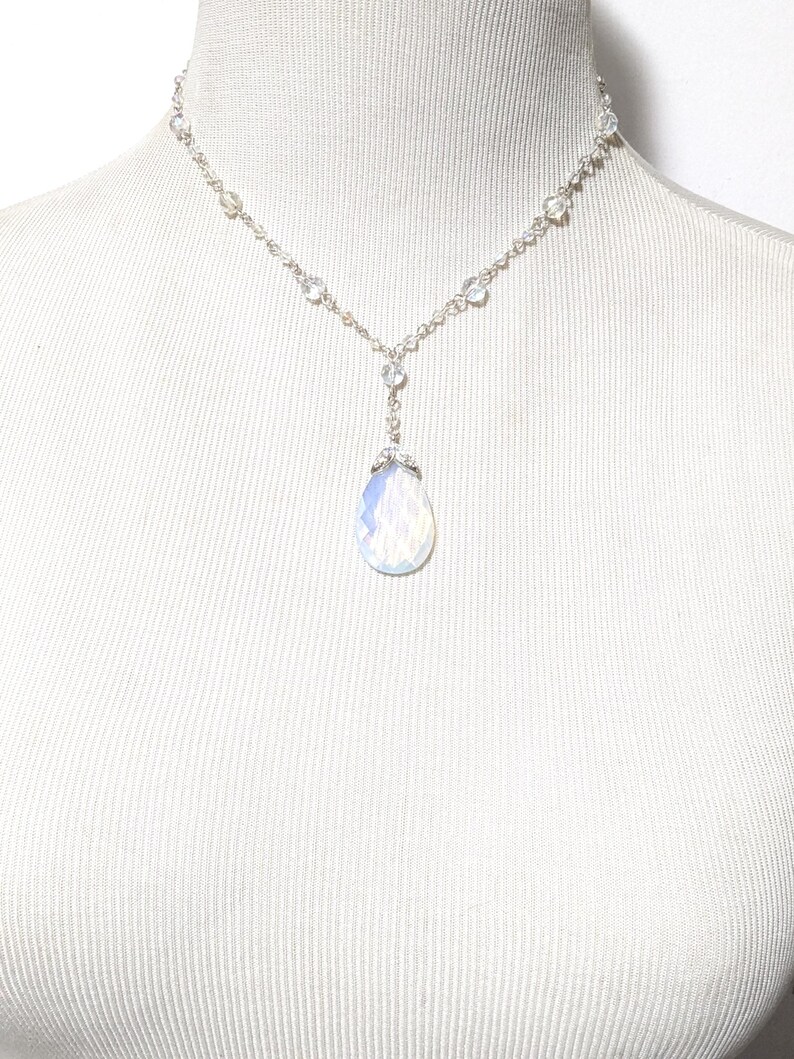 May include: A delicate silver necklace with a teardrop-shaped, iridescent pendant. The necklace features small, clear beads and a chain that drapes gracefully. The pendant is a soft, opalescent color, adding a touch of elegance.