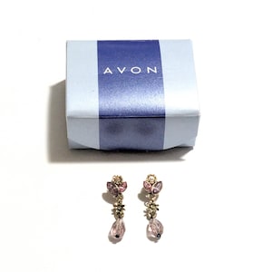 May include: A pair of gold-toned drop earrings with pink and purple accents, featuring floral and teardrop-shaped details. The earrings are displayed with a light blue box with the word "AVON" printed on it.